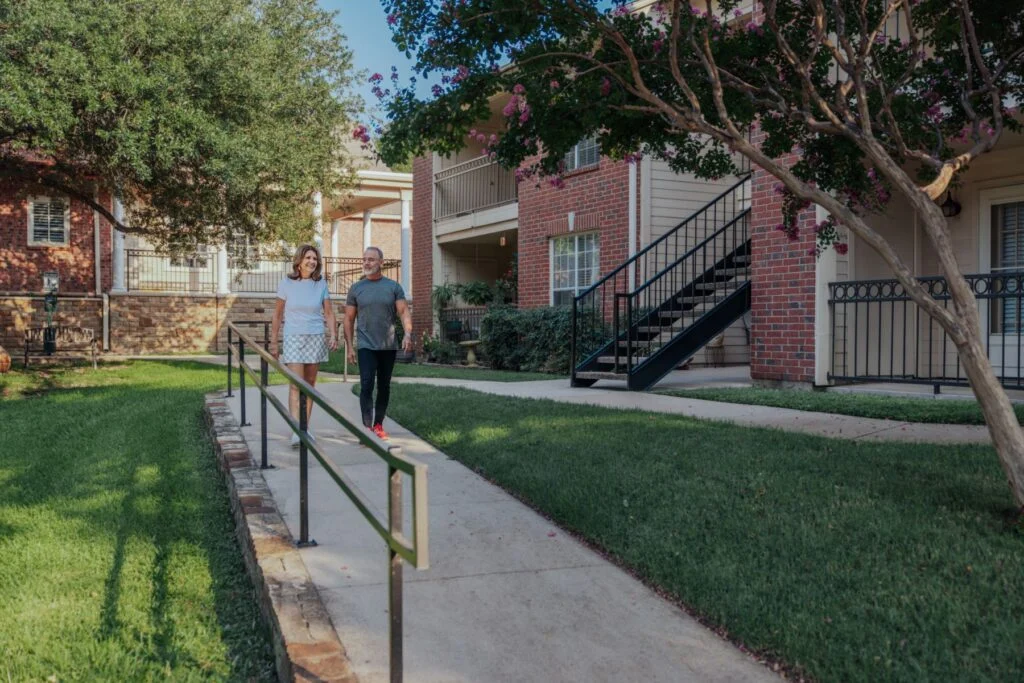 Two people walking along a sidewalk with a railing past a lawn and a brick apartment building