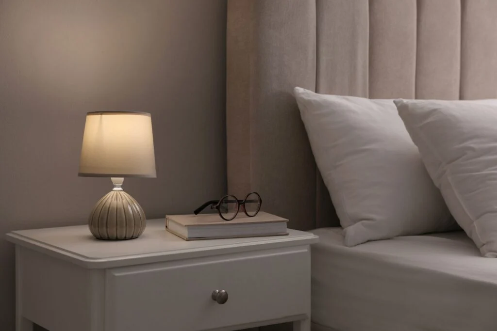 Bedside table with a lamp, glasses and a book beside a bed with white pillows