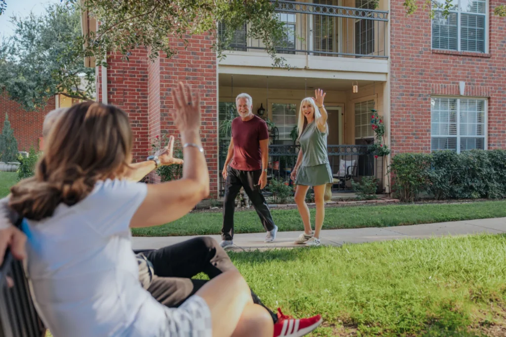 Woman seated on a bench waving to two friends walking across a lawn in front of a brick apartment building
