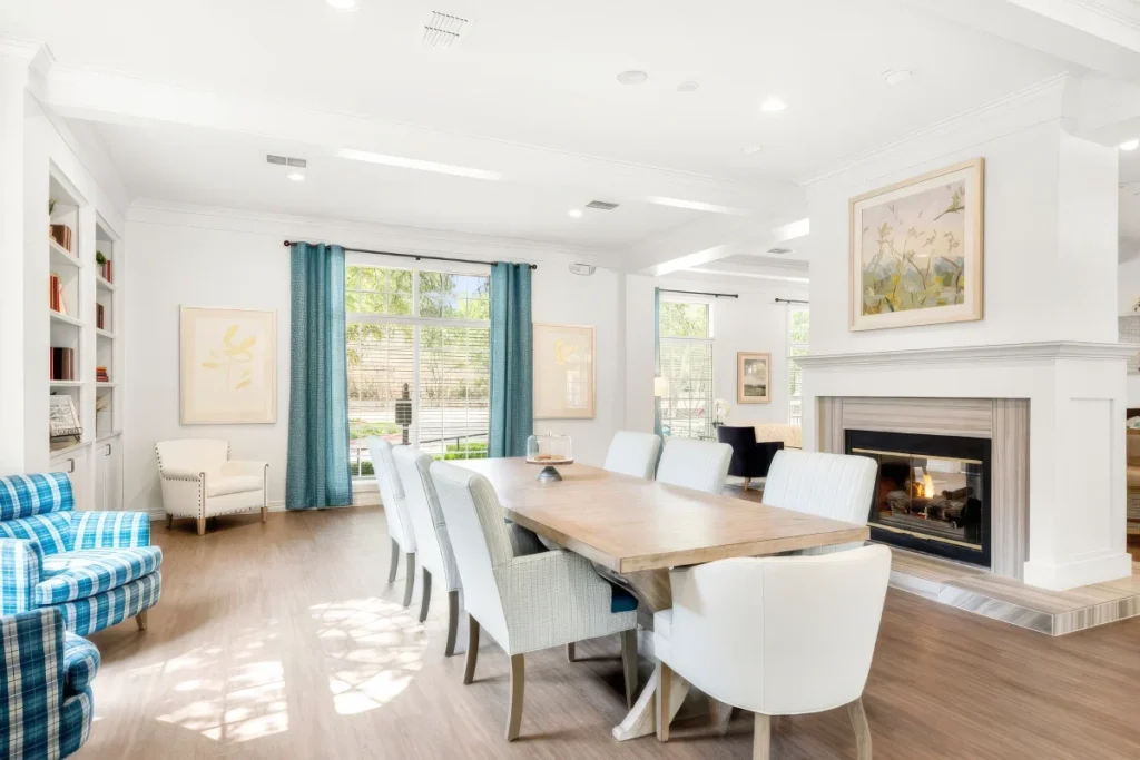 Bright common room with a long wooden dining table, white chairs and a fireplace, with teal curtains at large windows