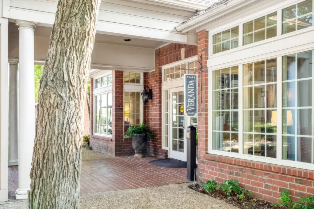 Covered brick building entrance with white columns and large windows, beside a tree trunk