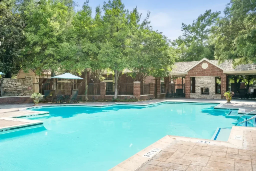 Outdoor swimming pool surrounded by trees, with a low brick building and umbrellas beyond