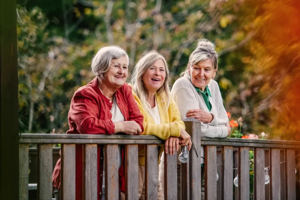 Three older women smiling together at a wooden railing among autumn trees